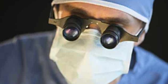 Surgeon wearing surgical loupes and a face mask, focused on a medical procedure. The image highlights the importance of precision in surgery, showcasing the tools that enhance visibility and accuracy during operations. The background is blurred, emphasizing the surgeon's concentration and professionalism in a clinical setting.